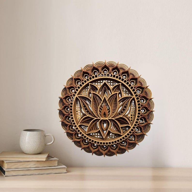 A layered wooden lotus mandala with light and dark wood tones hangs on a light beige wall above a wooden table with a white mug and books.