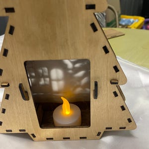 May include: A wooden Christmas tree shaped candle holder with a small white candle inside. The candle is lit and the flame is visible.