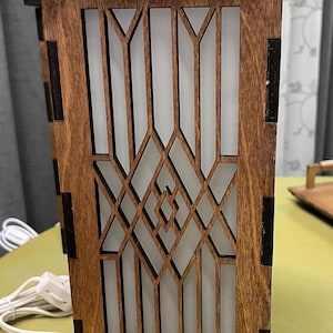 May include: A rectangular wooden table lamp featuring a laser-cut geometric lattice overlay in a dark brown finish. The structure houses a frosted white panel that diffuses light, with visible dark joinery details at the corners and a white power cord.
