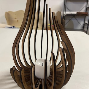 Op de afbeelding: Een houten kaarsenhouder met een uniek, organisch ontwerp. De houder is gemaakt van dunne stroken hout die gebogen en met elkaar verweven zijn om een ​​delicaat, luchtig uiterlijk te creëren. Een kleine witte kaars zit in de houder.