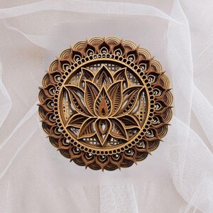 A layered wooden lotus mandala with light and dark wood tones is centered on a sheer white fabric background.