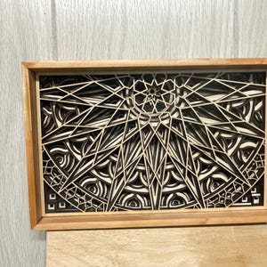 A rectangular wooden frame holds a layered mandala art piece with intricate geometric patterns in alternating light and dark wood tones.