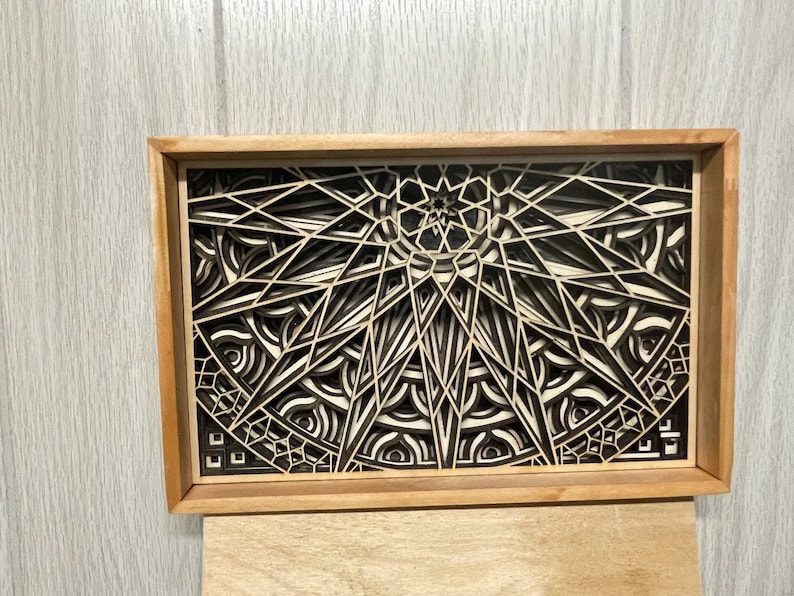 A rectangular wooden frame holds a layered mandala art piece with intricate geometric patterns in alternating light and dark wood tones.