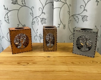 May include: Three wooden candle holders with a tree of life design. The candle holders are in shades of brown and gray, and are displayed on a wooden table. The tree of life design is cut out of the front of each holder.
