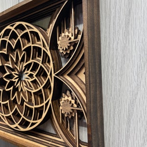 Puede incluir: Una pieza de arte mural de madera en capas con un marco marr&oacute;n oscuro, que presenta un dise&ntilde;o geom&eacute;trico. La obra de arte muestra un patr&oacute;n circular y acentos florales, creando un efecto tridimensional. La madera tiene un color marr&oacute;n claro natural.