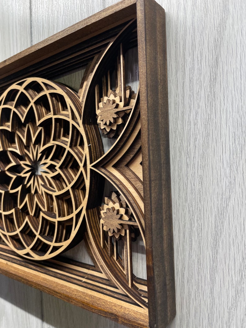 Puede incluir: Una pieza de arte mural de madera en capas con un marco marr&oacute;n oscuro, que presenta un dise&ntilde;o geom&eacute;trico. La obra de arte muestra un patr&oacute;n circular y acentos florales, creando un efecto tridimensional. La madera tiene un color marr&oacute;n claro natural.