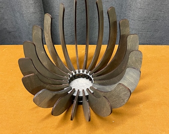 May include: A wooden decorative bowl with a flower-like design. The bowl is made of dark brown wood and has a circular base with curved, petal-shaped pieces that extend upwards. The interior base is a light gray color.