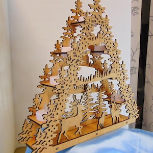 A laser-cut wooden Christmas decoration, angled to the right, shows a layered winter scene with deer, trees, and a house, with seven flameless tea lights.