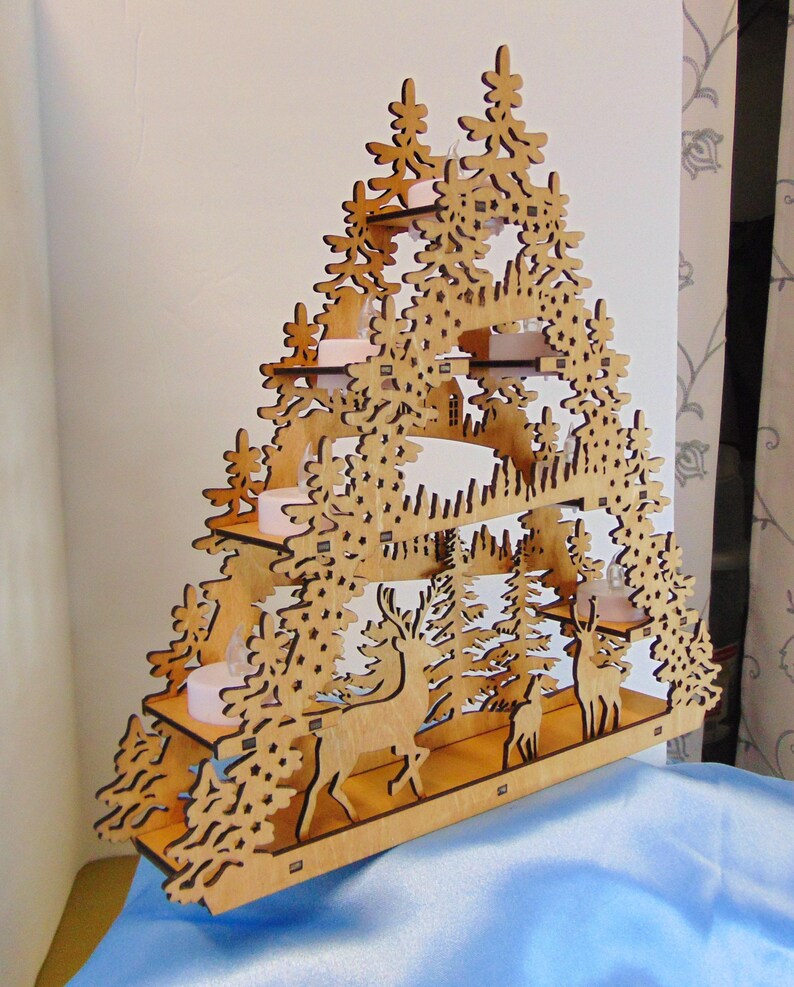 A laser-cut wooden Christmas decoration, angled to the right, shows a layered winter scene with deer, trees, and a house, with seven flameless tea lights.
