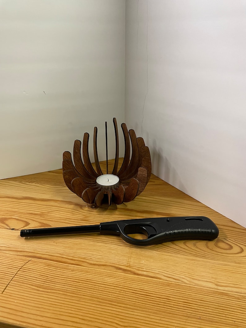 K&ouml;nnte beinhalten: Ein Kerzenhalter aus Holz in Form einer Blume mit einer kleinen wei&szlig;en Kerze darin. Ein schwarzer Butan-Feuerzeug liegt auf einer Holzoberfl&auml;che.