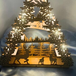 K&ouml;nnte beinhalten: Ein Weihnachtsbaum-Dekor aus Holz mit einem ausgeschnittenen Design einer Winterwald-Szene. Der Baum ist mit warmwei&szlig;en LED-Lichtern beleuchtet und zeigt Hirsch-Silhouetten.