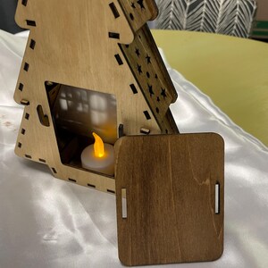 May include: A wooden Christmas tree shaped candle holder with a small tea light inside. The tree is made of light brown wood and has a dark brown stain on the back panel. The tree is decorated with stars and has a small door on the front.