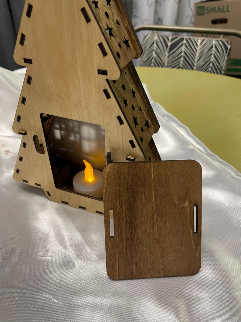 May include: A wooden Christmas tree shaped candle holder with a small tea light inside. The tree is made of light brown wood and has a dark brown stain on the back panel. The tree is decorated with stars and has a small door on the front.