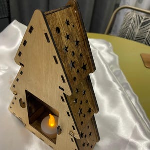 May include: A wooden Christmas tree shaped candle holder with a cutout design of stars. The holder has a small opening for a tea light candle. The candle holder is made of light brown wood with a darker brown stain.