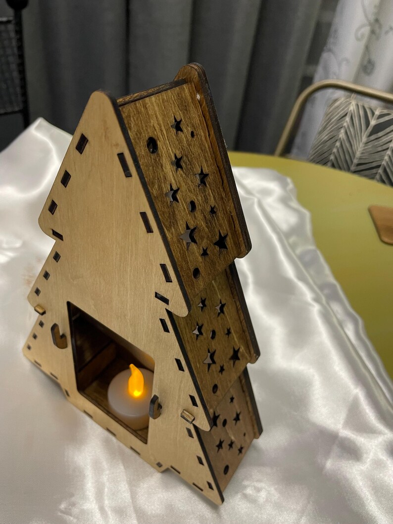 May include: A wooden Christmas tree shaped candle holder with a cutout design of stars. The holder has a small opening for a tea light candle. The candle holder is made of light brown wood with a darker brown stain.