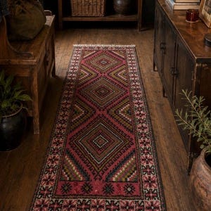 May include: A long rectangular runner rug featuring a deep pink background with a repeating pattern of large diamonds in shades of brown, green, and cream. The rug has a decorative border with small floral motifs and light-colored fringe at both ends.