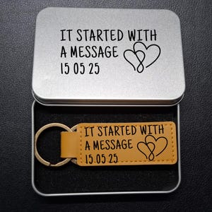 May include: A silver metal gift box with a hinged lid, open to reveal a tan leather keychain. The keychain and the inside of the lid both have the text "IT STARTED WITH A MESSAGE 15 05 25" and two intertwined hearts.