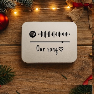 May include: A silver rectangular tin with the text "Our song" and a heart symbol. Above the text is a Spotify code and a graphic equalizer design. The tin is on a wooden surface with Christmas decorations.