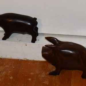 May include: Two dark brown wooden pig figurines. The pig on the left is standing on a white surface, while the pig on the right is standing on a wooden surface.