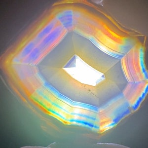 May include: A close-up of a colourful agate geode with iridescent bands of blue, green, yellow, and orange. The geode is cut in a square shape and has a hollow centre.