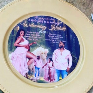May include: A yellow round plate with a white border and a printed design featuring a couple and a unicorn. The text on the plate reads "Once upon a time a little fairy is on her way Welcoming Kailani Menu". The menu includes items like chicken and waffles, cheese burger sliders, cinnamon French toast, shrimp and grits, home fried potatoes, applewood smoked bacon, turkey bacon, scrambled eggs with cheddar cheese, and fresh fruit.