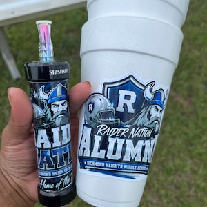 May include: A white foam cup and a cylindrical black plastic container, both featuring blue and white decals with a Viking mascot, a football helmet, and the text Raider Nation Alumni Richmond Heights Middle School and Home of the Raiders.