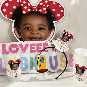 Może przedstawiać: Zestaw dekoracji na przyjęcie z motywem Minnie Mouse, przedstawiający uśmiechniętą twarz dziecka. Zawiera duży wycięty element, kubki, opaskę na głowę i znak z napisem "Loveee 1/2 Clubhouse". Kokarda Minnie Mouse jest czerwona w białe grochy.