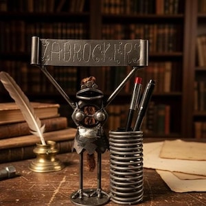 Może przedstawiać: Metalowy organizer na biurko w kształcie żaby trzymającej znak z napisem "ZABROCKERS". Żaba ma sprężynowy uchwyt na długopisy i trzyma długopisy. Na biurku znajdują się również pióro i kałamarz.