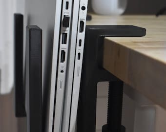 May include: A black metal laptop holder mounted to a wooden desk. Two silver laptops are held vertically in the holder. The holder is clamped to the desk with a screw mechanism. A white ceramic cup with pens is in the background.