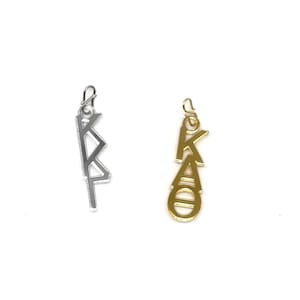 May include: Two metal charms, one silver and one gold, each featuring Greek letters. The silver charm displays the letters "KAP" in a stylized, angular design. The gold charm shows the letters "KAO" in a more rounded, connected font. Both charms have a small loop at the top.