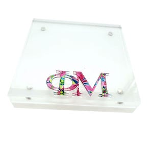May include: A clear, square acrylic block featuring the Greek letters Phi and Mu in a vibrant, multi-colored floral pattern. The letters are mounted on the surface of the transparent block, which is secured with four small silver-toned metal screws.
