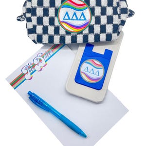May include: A white notepad with a rainbow stripe design and the text "Tri Delta" in a retro font. A blue pen, a white phone case with a blue pop socket featuring a rainbow and the letters "AAA", and a black and white checkered pouch with a rainbow and the letters "AAA" are also included.