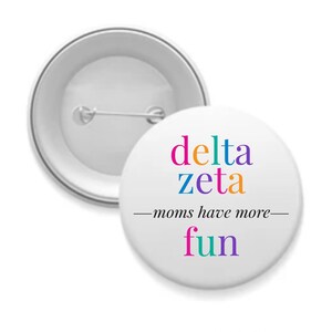 May include: A white button with a silver border and a pin on the back. The front of the button has the text "delta zeta - moms have more fun" in colorful letters.