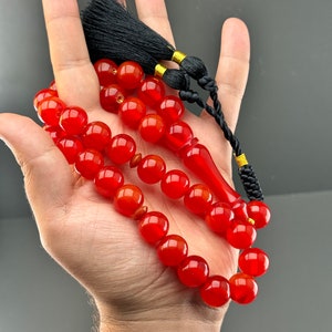 May include: A red carnelian prayer bead mala with a black tassel and gold accents. The beads are round and smooth, and the tassel is long and flowing.