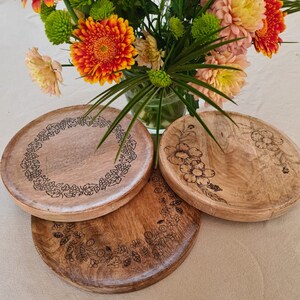 May include: Three round wooden serving trays with intricate floral designs burned into the surface. The trays are stacked on top of each other.
