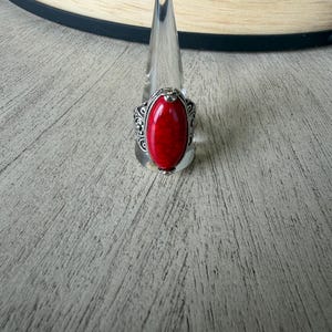 May include: A silver-toned ring featuring an oval-shaped, bright red stone with dark veining. The metal band displays intricate, embossed scrollwork patterns along the sides, framing the central cabochon stone in a secure, polished setting.