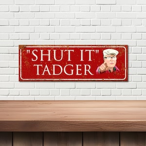 May include: Red metal sign with a distressed look featuring a cartoon man with a white cap and glasses pointing at the viewer. The sign reads "SHUT IT TADGER".
