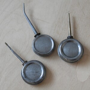 May include: Three antique metal oil cans with spouts of different lengths. The cans are circular with a silver-grey finish, showing signs of wear. The spouts are thin and metallic, with one curved and two straight.