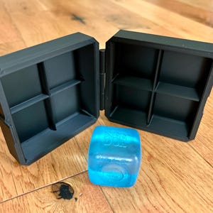 May include: A black, hinged, square storage case with two open compartments, each divided into four smaller sections. Beside the case sits a translucent, bright blue cube with the embossed text "LIFE DIE" visible on its top surface.