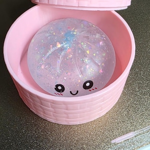 May include: A round, light pink container with a textured basket-weave pattern and a matching lid. Inside sits a clear, squishy, spherical toy filled with iridescent glitter and featuring a printed face with black eyes and a smiling mouth.