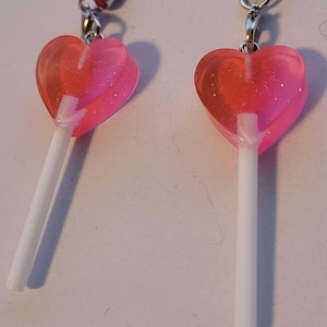 Könnte beinhalten: Ein Paar rosa herzförmige Lollipop-Ohrringe mit silbernen Haken und weißen Stielen.