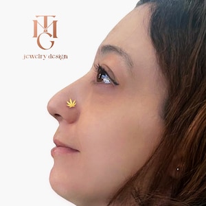 May include: A close-up of a person's face with a gold marijuana leaf nose ring. The person is looking to the side and has dark hair.