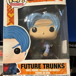 Puede incluir: Una figura de vinilo Funko Pop! de Future Trunks de Dragon Ball Super, de color azul y blanco. La figura está en una caja de plástico transparente con un fondo amarillo y naranja. La caja tiene las palabras "Pop! Animation" y "Dragon Ball Super" en ella. La figura está numerada 313.