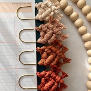 May include: Four handmade macrame bookmarks in shades of beige, peach, orange, and red. Each bookmark is attached to a gold metal bookmark clip. The bookmarks are arranged vertically on a teal background.