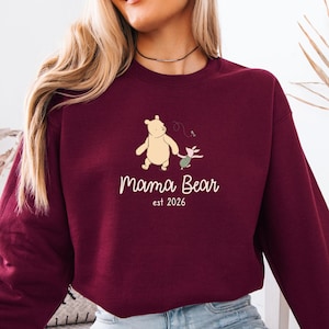 Op de afbeelding: Bordeaux sweatshirt met een cartoon beer en varken illustratie. De tekst "Mama Bear est 2026" is onder de illustratie gedrukt in een cursief lettertype. De sweatshirt is effen van kleur.