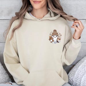 May include: Beige hoodie with a front pocket and a drawstring hood. The hoodie features a small graphic of a ghost wearing a knit hat, holding a teddy bear and a cupcake. The graphic is in shades of brown and white.