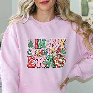 May include: A light pink sweatshirt with the text "IN MY CHRISTMAS ERA" in colorful, retro-style lettering. The design includes a Christmas tree, disco ball, and festive lights, perfect for the holiday season.