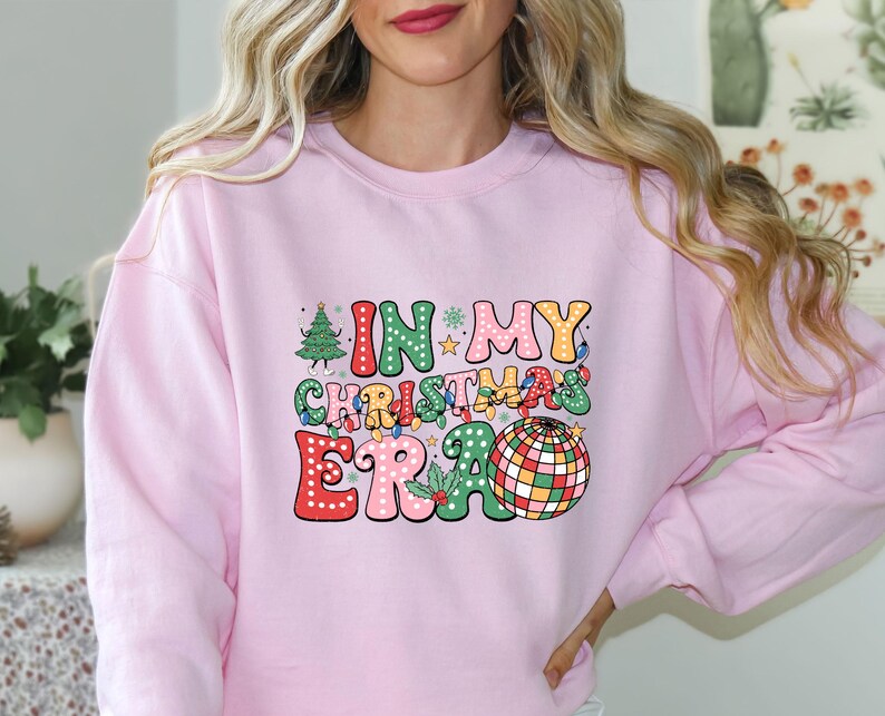 May include: A light pink sweatshirt with the text "IN MY CHRISTMAS ERA" in colorful, retro-style lettering. The design includes a Christmas tree, disco ball, and festive lights, perfect for the holiday season.