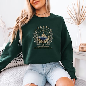 May include: A dark green long-sleeve sweatshirt featuring a gold and blue graphic print of mountains and stars. The text reads "VELARIS CITY OF STARLIGHT" and "To the stars who listen and the dreams that are answered" encircled by a leafy laurel wreath.