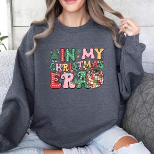 May include: Dark gray sweatshirt with the text "IN MY CHRISTMAS ERA" in colorful, retro-style lettering. The design includes a Christmas tree, a disco ball, and other festive elements, perfect for the holiday season.